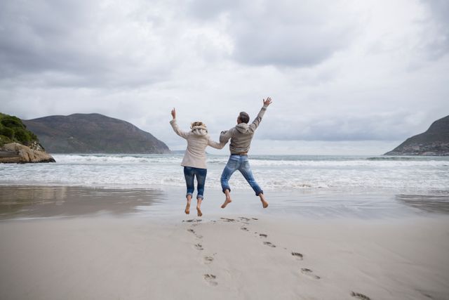 Mature couple jumping in the air on a beach, showing joy and togetherness. Ideal for use in travel brochures, vacation advertisements, lifestyle blogs, and wellness campaigns. Highlights themes of happiness, leisure, and active living.