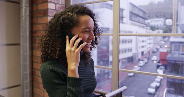Young woman with curly hair smiling while talking on phone in modern office with city view through the window. Ideal for depicting professional communications, business environment, positive workplace atmosphere, or corporate settings.