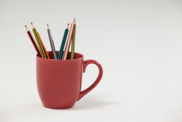 Colored pencils in a red cup on a white background. Ideal for use in educational materials, office supply advertisements, or creative art projects. The minimalistic and clean design makes it suitable for various themes related to creativity, organization, and learning.