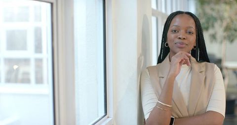 African American woman leaning by office window resting chin wearing blazer and smartwatch