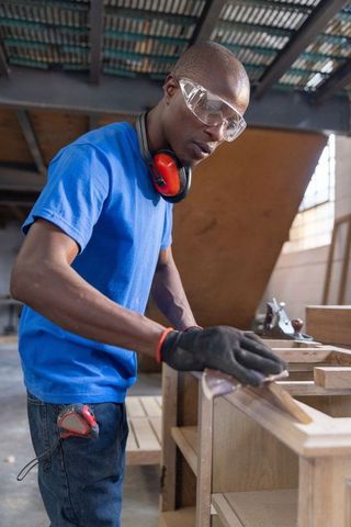 African american carpenter sanding wood at workbench in workshop wearing safety gear