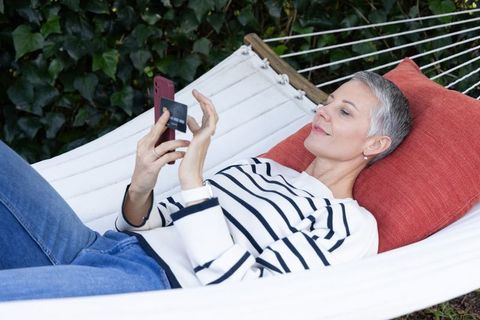 Relaxing mature woman making contactless payment on phone in backyard hammock