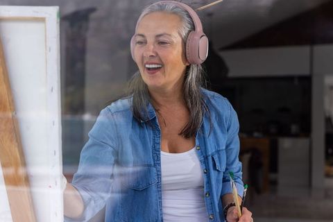 Senior woman painting at easel wearing headphones in bright home art studio