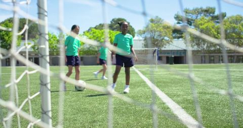 Diverse teenage girls practicing soccer on school turf through goal net during training