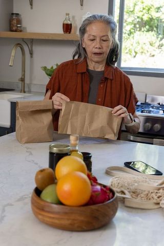 Senior asian woman folding paper bag on marble kitchen island with wooden fruit bowl meal prep