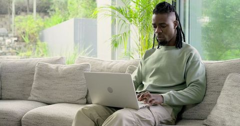 African American man working on laptop on modern sunlit sofa with indoor plants