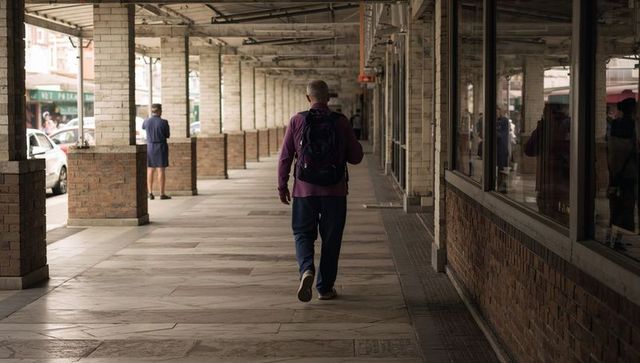 Senior man walking under covered arcade carrying backpack wearing maroon jacket and pants