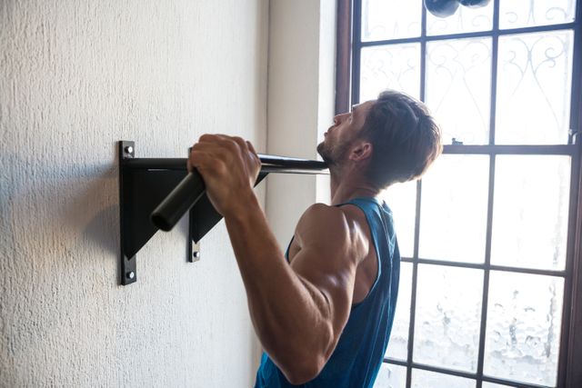 Side view of male athlete practicing pull ups on bar by window at fitness studio