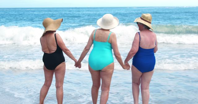 Three senior women standing on the shore, holding hands and enjoying the beach atmosphere. Ideal for concepts of friendship, aging gracefully, summer vacations, relaxation by the ocean, and spending quality time together.