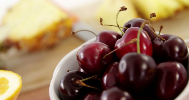 A bowl of ripe cherries is in the foreground, with a blurred background featuring slices of citrus and cake, with copy space. Fresh cherries are often associated with summer and can be a healthy, sweet addition to any meal or snack time.