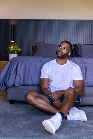 African american man sitting on floor by bed, looking thoughtful in minimalist bedroom