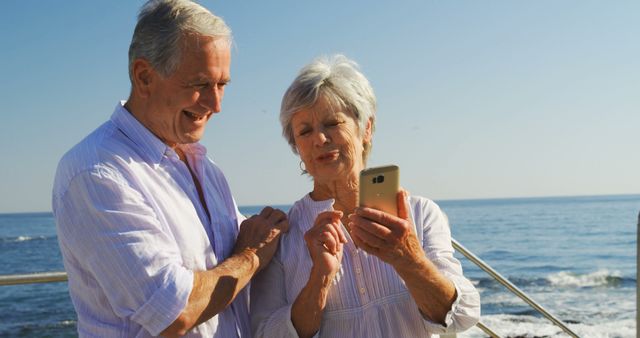 Happy elderly couple stands by the ocean, smiling while reviewing photos on a smartphone. Perfect for illustrating themes like technology for seniors, vacation, retired life, and happiness. Ideal for brochures on senior travel, advertisements for smartphones, or lifestyle articles.