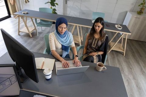 Multicultural women collaborating on laptop in modern coworking office, wearing hijab and ethnic dre