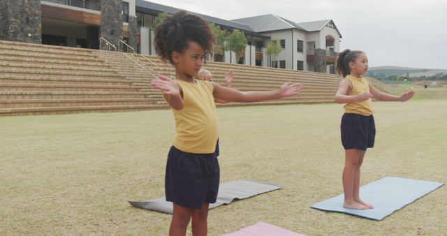 Three young girls practicing yoga outdoors on a cloudy day. They are standing on yoga mats in a park, focusing and stretching. This image can be used in materials promoting children's health, fitness programs, summer camps, or mindfulness activities targeted at young kids.