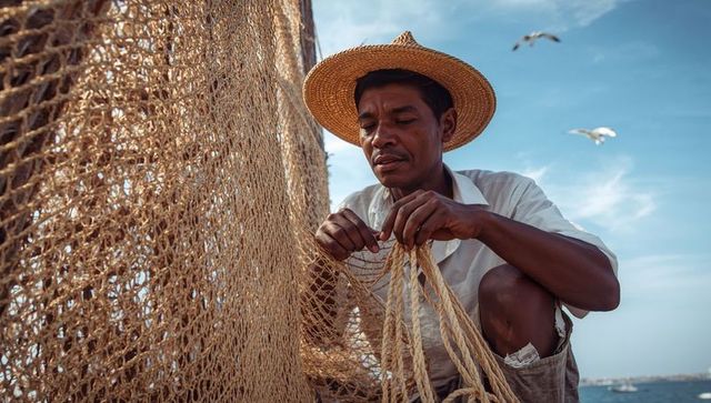 Sunlit coastal fisherman mending woven fishing net and ropes on boat deck wearing straw hat