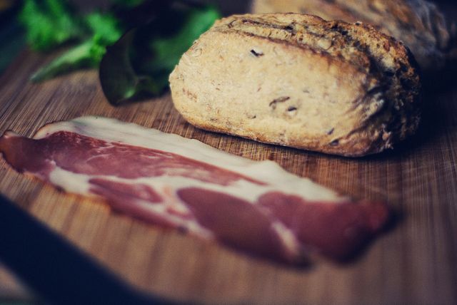 Rustic bread placed next to thin slices of prosciutto on a wooden board with leafy greens. Great for use in culinary blogs, recipe websites, or gourmet food advertisements. Perfect for illustrating charcuterie preparation, sandwich making, or highlighting rustic, homecooked meals.