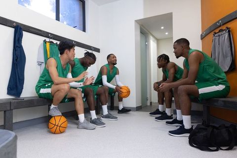 Diverse male basketball team sitting and bonding in locker room holding basketballs