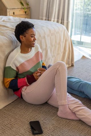Smiling african american woman relaxing on bedroom floor holding mug, striped sweater
