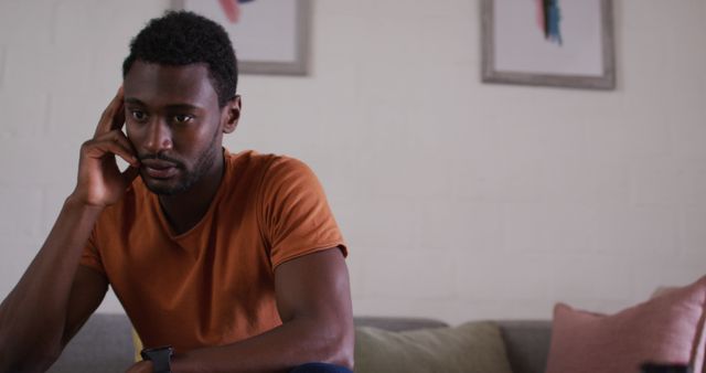 Young man wearing orange T-shirt and watch, seated on a couch in modern living room, looking thoughtfully at phone, suitable for themes of communication, technology, home life, and contemplative moments.