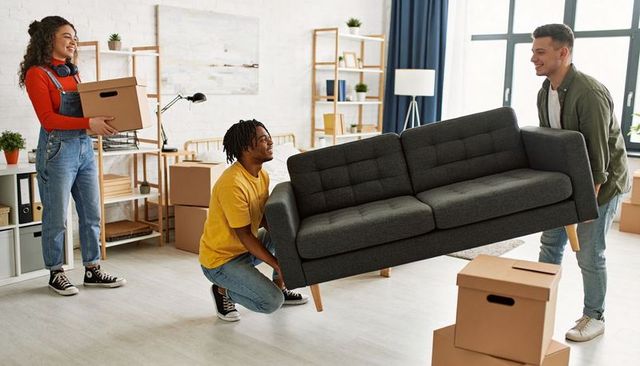 Friends moving into new apartment carrying dark gray sofa and boxes, teamwork