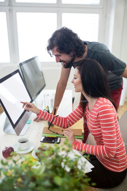 A male and female graphic designer are engaging in a creative discussion over a computer in a modern office. This scene highlights collaboration, teamwork, and creativity in a professional setting. Perfect for illustrating concepts related to creative work environment, design processes, and professional collaboration.