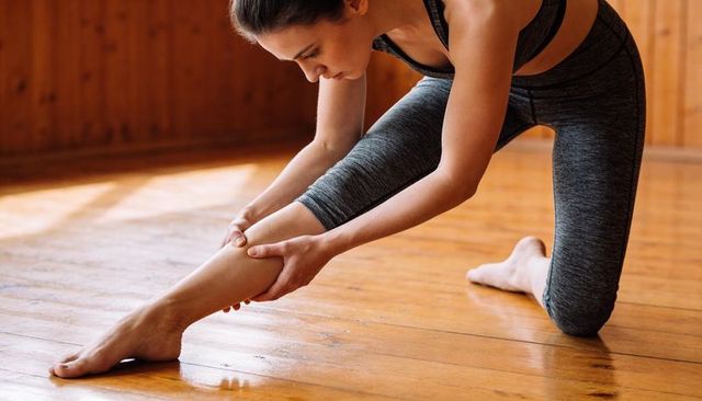 Kneeling athlete stretching calf in sunlit wooden studio wearing grey tights and bra