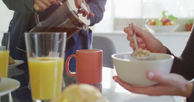 Cozy morning breakfast scene featuring two people enjoying coffee, cereal, and orange juice at a kitchen table. One is pouring coffee into a mug while another is holding a bowl of cereal. Ideal for use in lifestyle blogs, healthy eating promotions, kitchen and home decor advertisements, and food and beverage marketing materials.