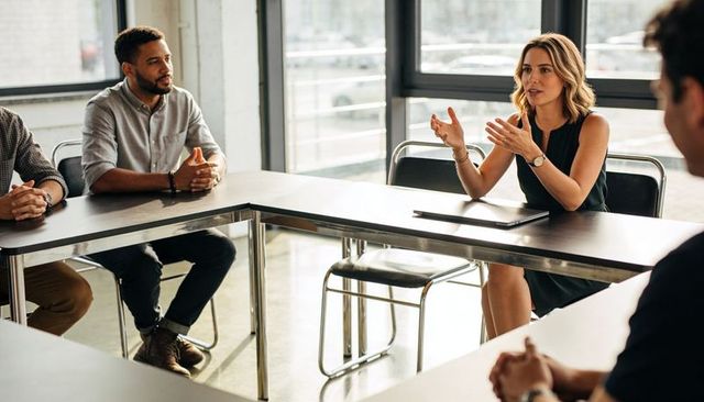 Female leader leading meeting and gesturing in modern sunlit office with collaborative team