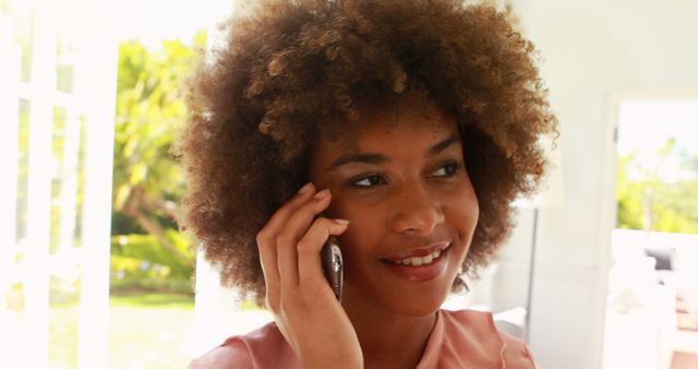Young Afro-American woman with afro hair is happily talking on a smartphone in a bright indoor space with natural light. This image can be used for business communications, customer service, social media campaigns, technology advertisements, and lifestyle blogs.