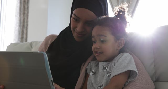 Mother and daughter are sitting together on a couch, reading from a tablet. The mother wears a hijab, indicating cultural background, and they are enjoying quality family time. This can be used in contexts promoting family bonding, technology in everyday life, parenting, and multicultural representation.