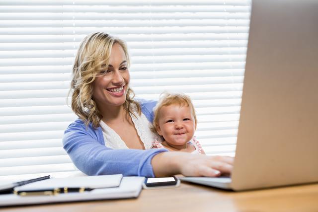 Mother holding baby girl on lap while using laptop in bright, cozy home environment. Ideal for themes related to work-life balance, parenthood, remote work, family bonding, and modern technology in everyday life.