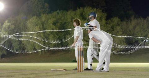 Night cricket trio standing at crease under floodlights with helmeted batsman and stumps