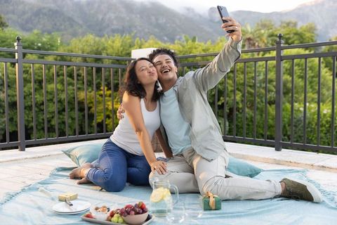 Romantic couple enjoying rooftop picnic at golden hour taking selfie with mountain view