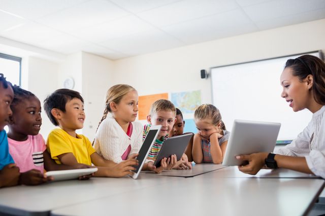 Female teacher using tablet to teach group of diverse students in classroom. Children engaged and interacting with digital devices. Suitable image for educational articles, school brochures, technology in education content, and advertisements promoting innovative teaching methods.