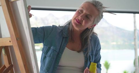Senior woman painting on canvas by window wearing denim shirt at easel