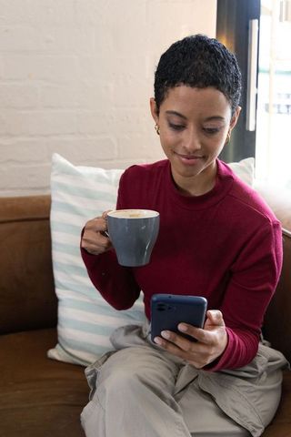 African american woman sipping coffee and checking smartphone on cozy sofa