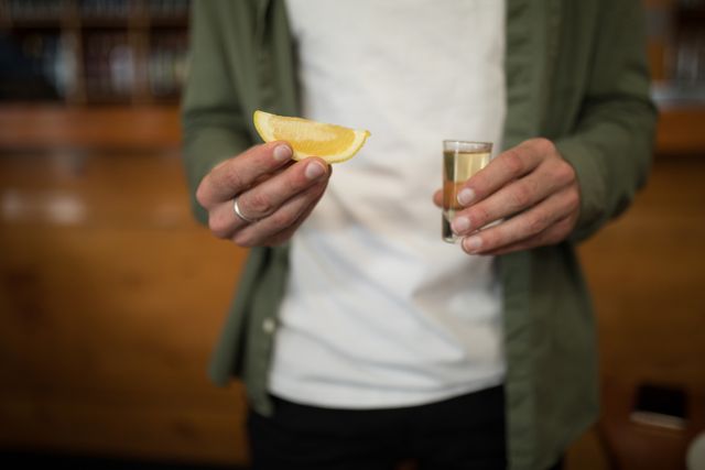 Man holding a tequila shot and lemon wedge in a bar, perfect for illustrating nightlife, social events, or casual drinking scenes. Useful for advertisements, blog posts about bar culture, or social media content related to parties and celebrations.