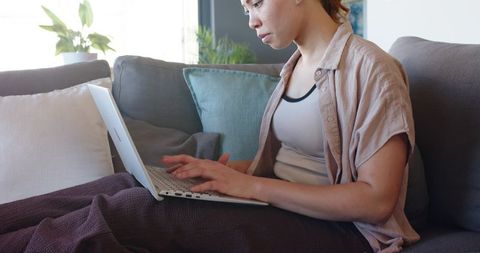 Young woman working on laptop on couch with blanket in daylight, cozy remote work