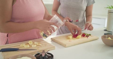 Women preparing fresh fruit smoothies in bright kitchen cutting berries and slicing banana