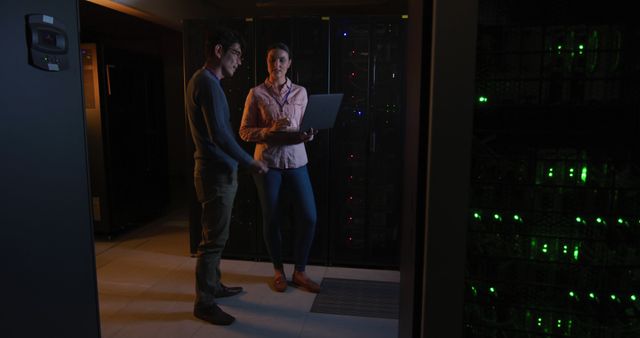 Two IT professionals collaborate in a dimly lit data center, standing near server racks and discussing information displayed on a laptop. The scene emphasizes technology, teamwork, and a high-tech work environment suited for promoting IT solutions, tech-driven teamwork, and efficient network system maintenance.