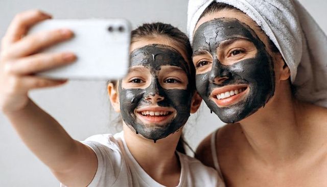 Smiling mother and daughter taking selfie while wearing charcoal facial masks and towel-wrapped hair