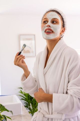 Woman applying facial mask with brush in white robe for at-home spa morning self-care