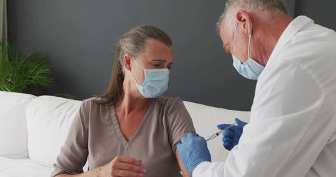 Home vaccination: Clinician administering vaccine to mature woman on sofa wearing masks