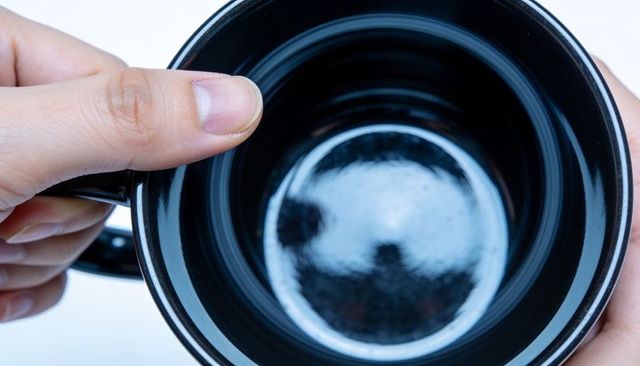 Top-down close-up hands holding glossy black ceramic mug with bright circular reflection