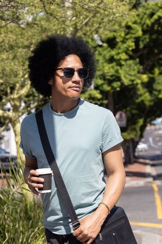 African american man walking with coffee and messenger bag on sunny urban sidewalk