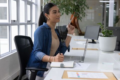 Young professional woman writing in notebook at bright open-plan office desk with laptop