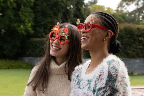 Diverse women wearing festive novelty glasses and cozy knit sweaters smiling outdoors