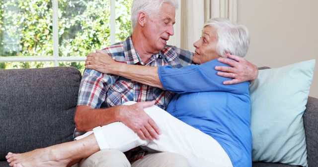 Senior man and woman sit hugging on couch in bright living room. Ideal for depicting senior romance, family warmth, and happily aging themes in marketing, healthcare advertisements, or elder care promotion.