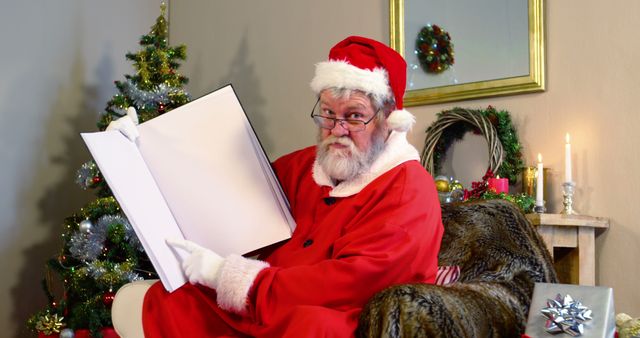 A Caucasian senior man dressed as Santa Claus looks surprised as he opens a large blank book, with copy space. His costume and the Christmas tree in the background set a festive holiday mood.