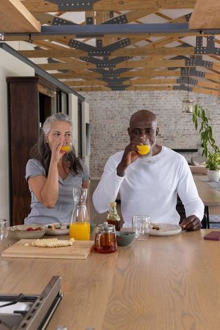 Mature diverse couple sipping orange juice sharing cozy breakfast at rustic kitchen island